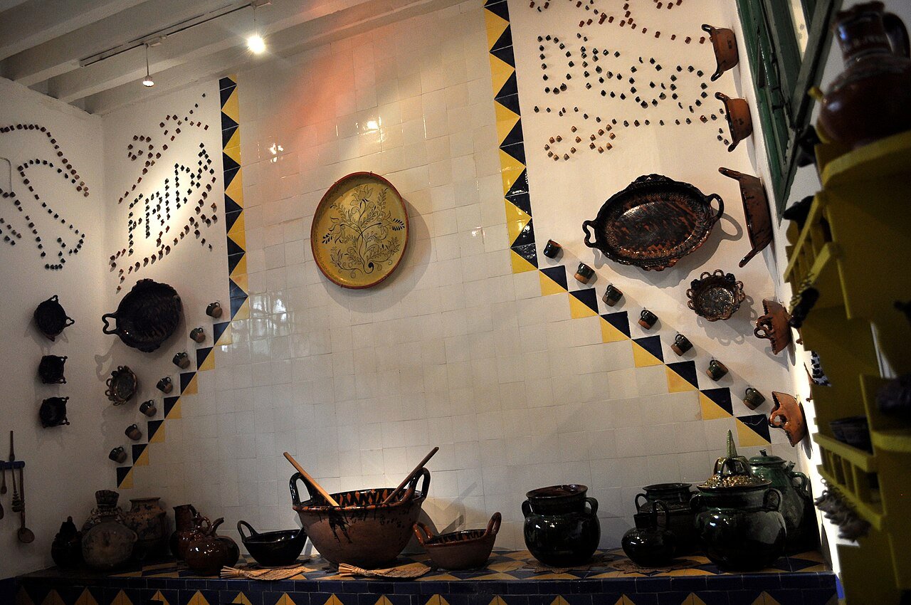 Parede de azulejos brancos com os nomes Frida e Diego formados por pequenas peças de barro, faixa de azulejos azuis e amarelos em zigue-zague, prato decorativo na parede e panelas e jarros de barro sobre a bancada