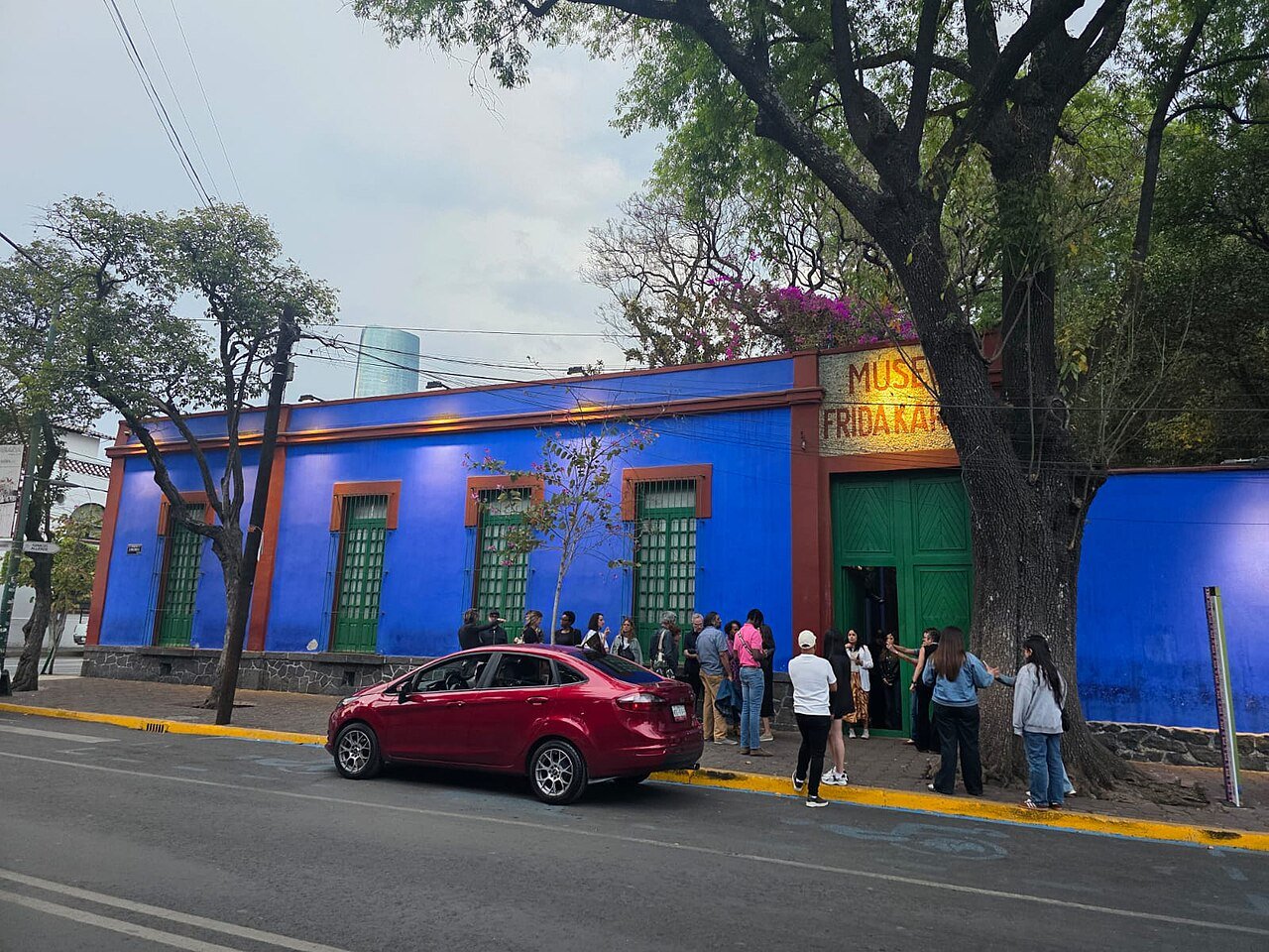 Fachada azul-cobalto da Casa Azul, com janelas de venezianas verdes, portão verde, árvores na calçada e visitantes na entrada do museu