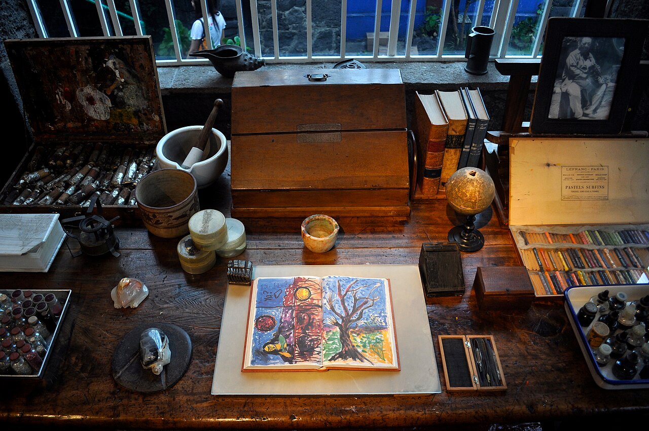 Mesa de madeira escura com paleta de tintas, caixa de pastéis, almofariz, livros, conchas e um caderno aberto com desenho colorido, com janela de grade ao fundo