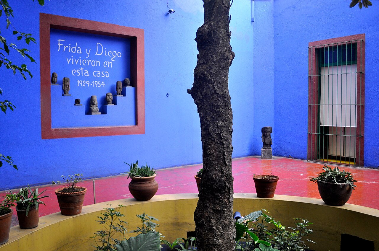 Pátio da Casa Azul com parede azul-cobalto, placa com os dizeres Frida y Diego vivieron en esta casa 1929-1954, piso vermelho e vasos de plantas ao redor de um tronco