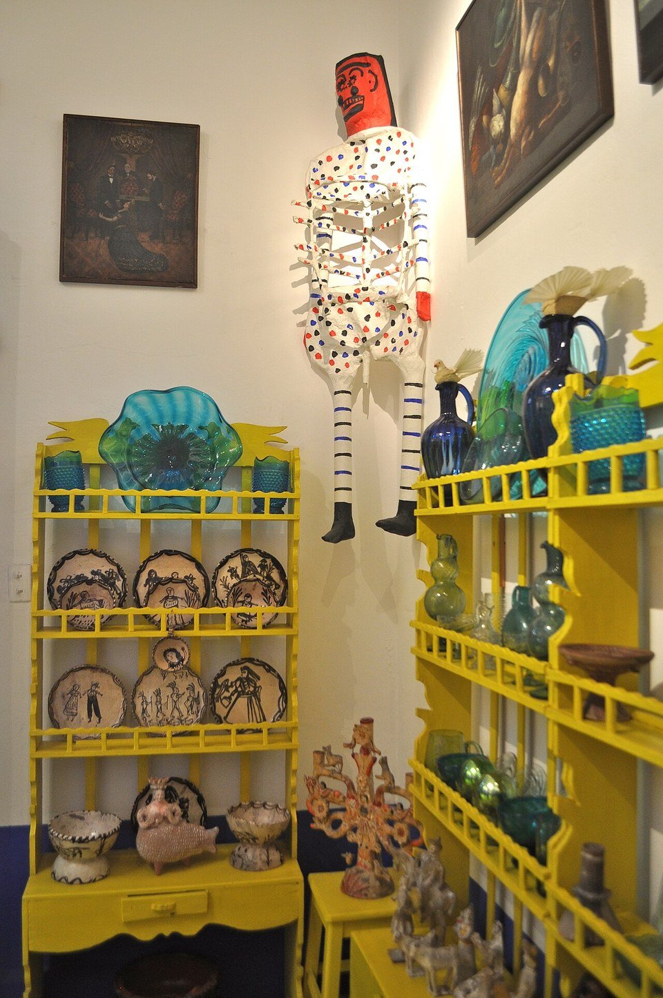 Duas estantes pintadas de amarelo com pratos decorados, jarras e vasos de vidro azul-turquesa, peças de cerâmica e uma figura de esqueleto articulada pendurada na parede branca, ao lado de quadros