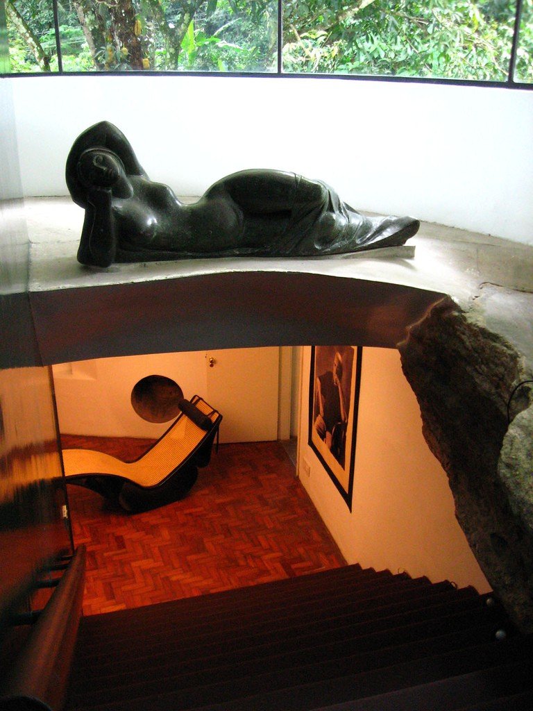 Vista de cima para baixo: no alto, uma escultura reclinada sobre a laje curva; embaixo, uma chaise de formas fluidas em um ambiente de piso vermelho, ao lado de uma grande pedra