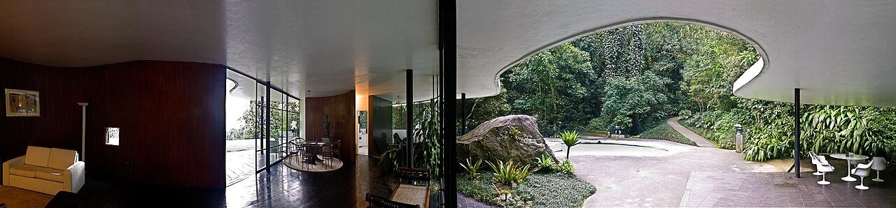 Panorama do interior da Casa das Canoas: à esquerda, um ambiente escuro com sofá claro; à direita, uma grande pedra e uma abertura curva para a piscina e a vegetação