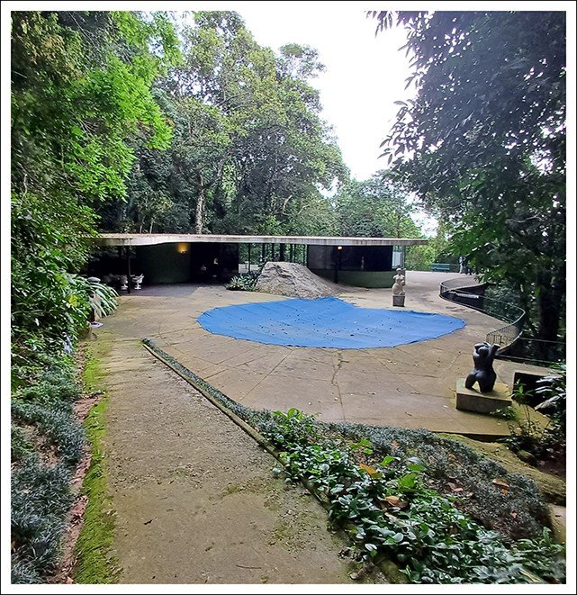 Caminho de terra que leva a uma piscina de contorno irregular coberta por lona azul, uma pedra e esculturas, com o pavilhão da casa ao fundo entre árvores