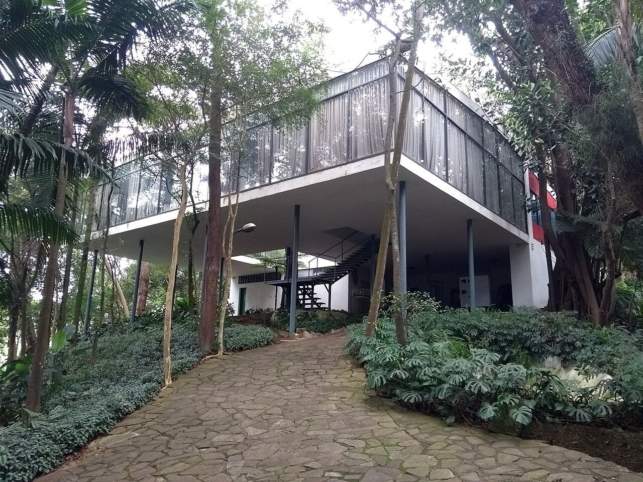 Casa de Vidro vista de um caminho de pedras cercado de vegetação densa, com o volume de vidro elevado sobre pilotis e escada metálica ao fundo