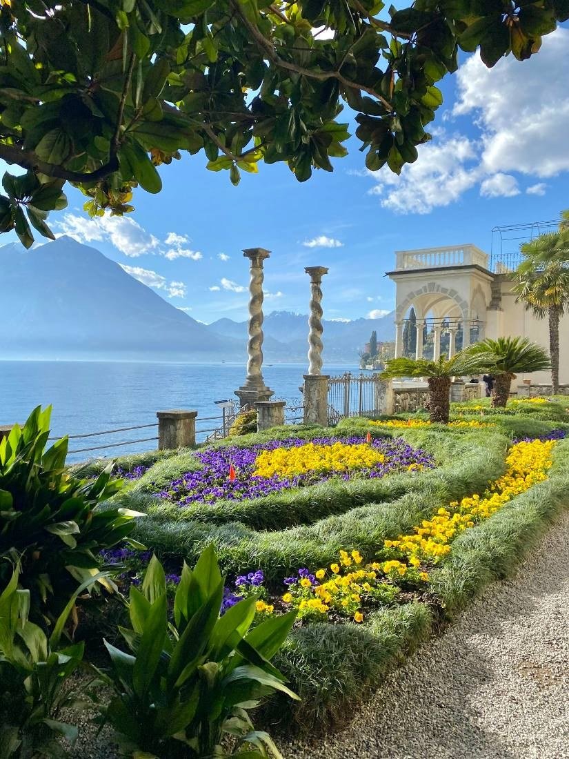 Canteiros com flores amarelas e roxas, colunas de pedra e o lago ao fundo, vistos sob galhos de uma árvore