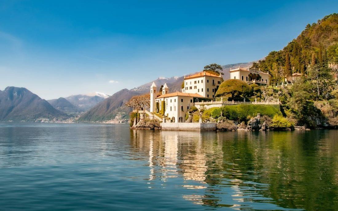 Vila histórica de paredes claras e telhado de telhas à beira da água do Lago de Como, com montanhas ao fundo