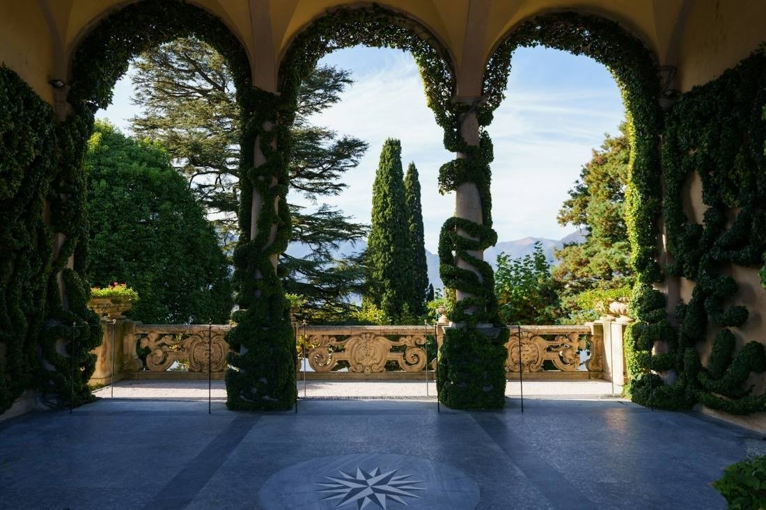 Terraço com três arcos cobertos de folhagem, balaustrada de pedra e vista do lago e de montanhas ao fundo