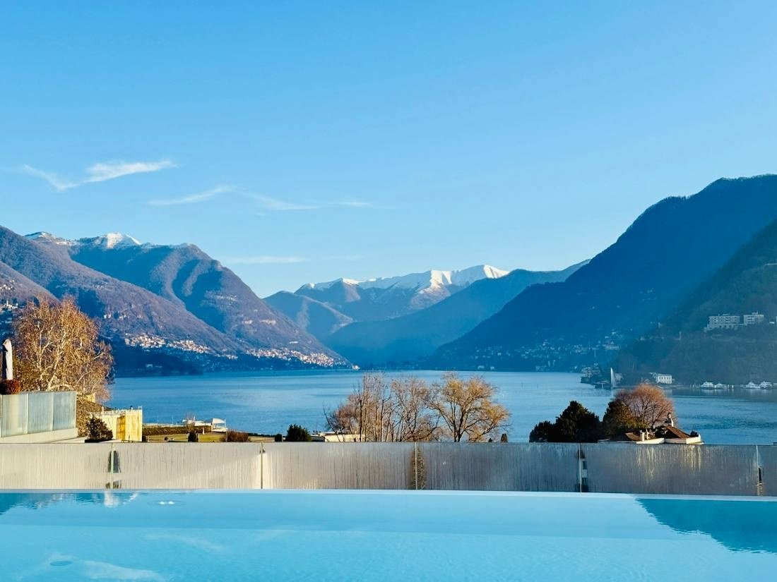 Borda de piscina em primeiro plano, com o lago e montanhas de topo nevado ao fundo sob céu azul