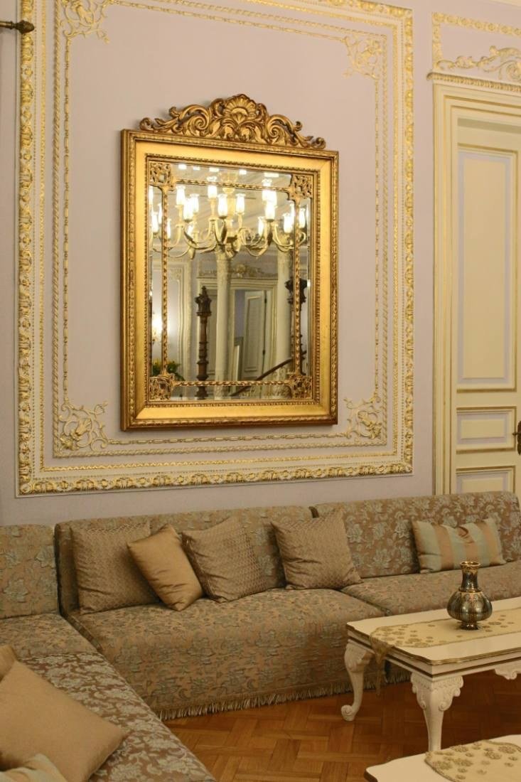 Sala clássica com boiserie em tom claro, espelho com moldura dourada e sofá com almofadas em tons de areia