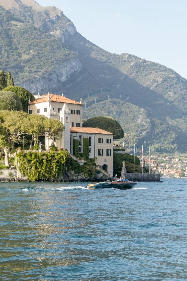 Casa de paredes claras e telhado de telhas entre árvores, vista de um barco no Lago de Como, com montanha ao fundo