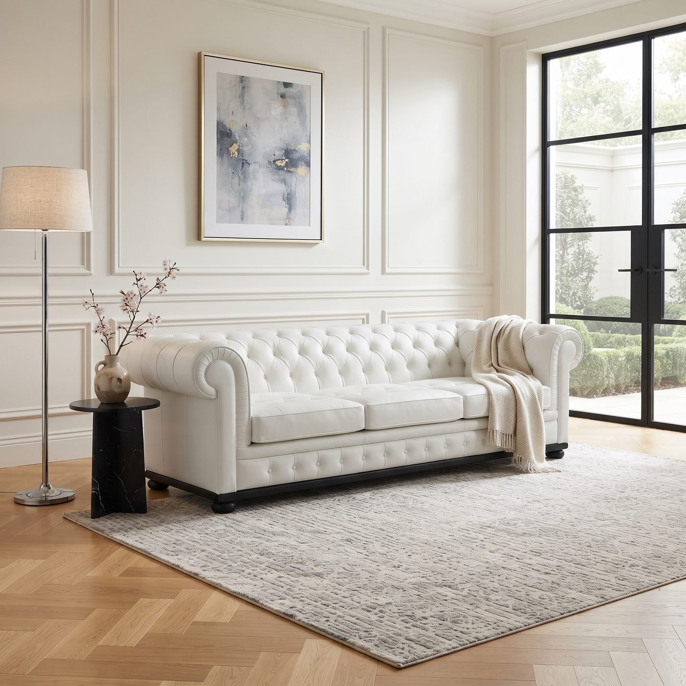 Estofado Chesterfield (Modelo A) branco em sala de paredes almofadadas claras, com manta sobre o braco, quadro abstrato acima e tapete claro no piso de madeira
